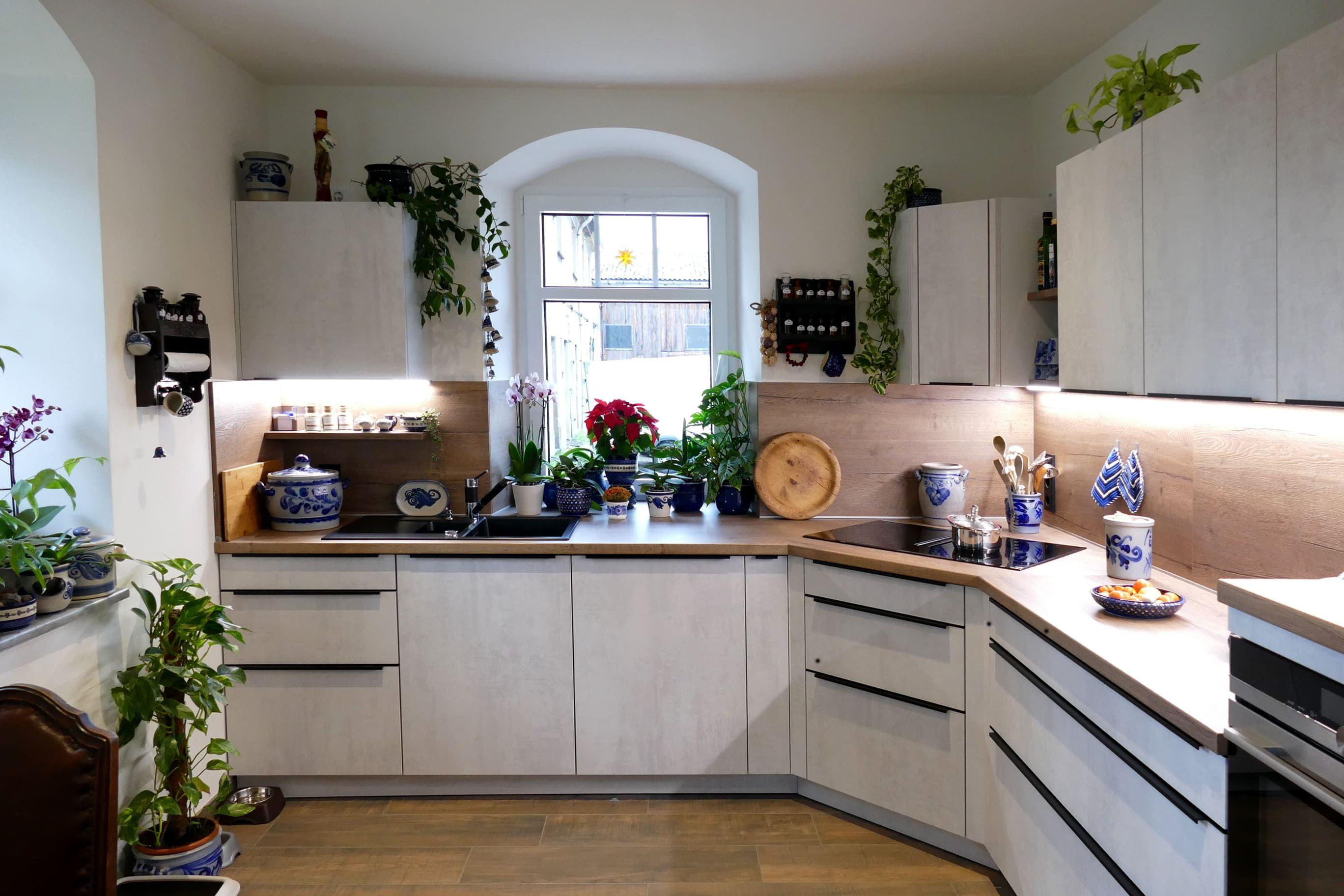 Helle Küche mit weißen Fronten, Holzarbeitsplatte und vielen Pflanzen rund um das Fenster, geplant vom Küchenstudio Hölzel Küchen Design.