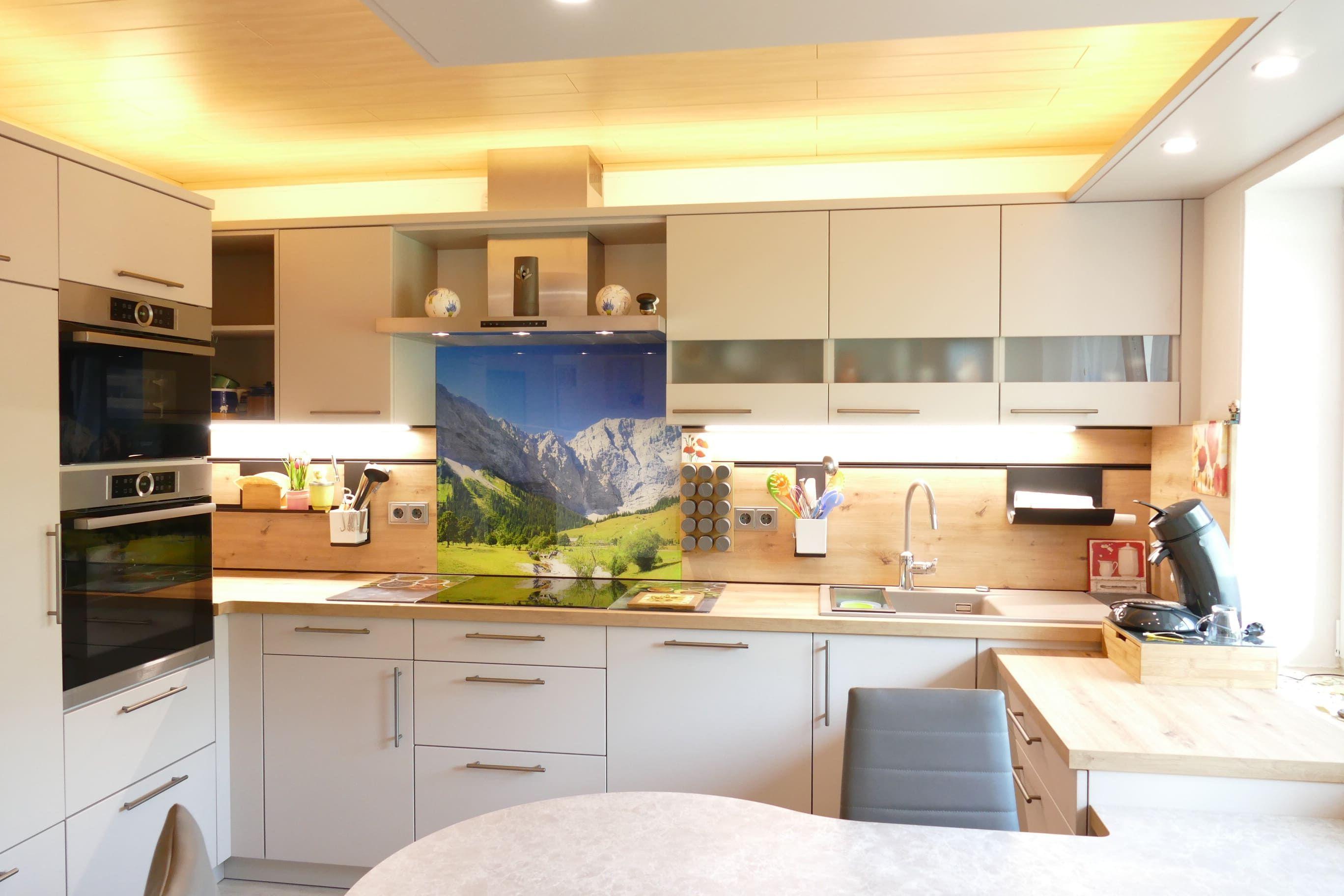 Helle Küche mit großem Bergmotiv als Rückwand und indirekter Beleuchtung, geplant vom Küchenstudio Hölzel Küchen Design.