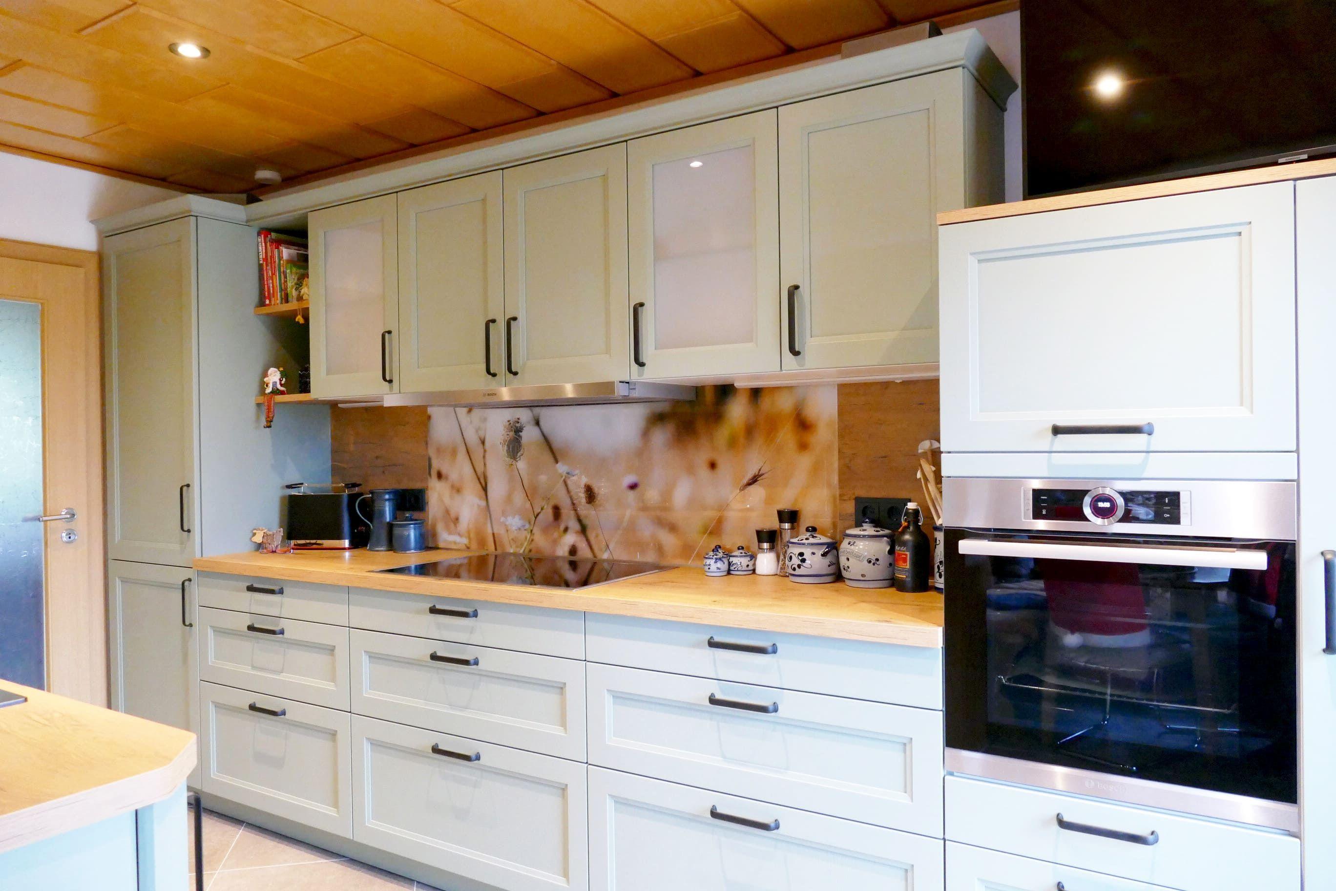 Landhausküche mit grün lackierten Rahmenfronten, Holzarbeitsplatte und dekorativer Rückwand, geplant vom Küchenstudio Hölzel Küchen Design.