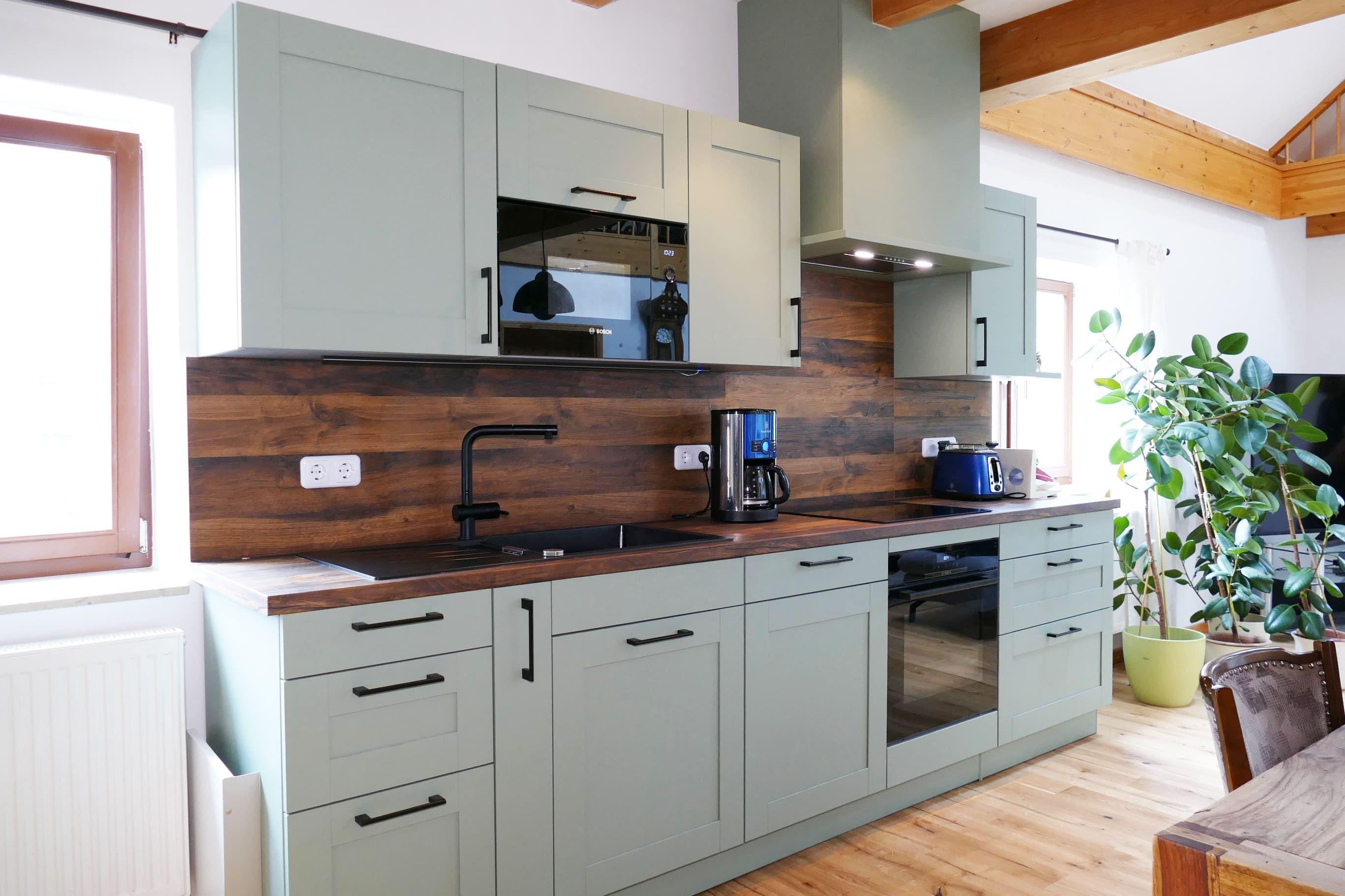 Moderne Küche mit salbeigrünen Fronten, dunkler Holzrückwand und schwarzem Wasserhahn, geplant vom Küchenstudio Hölzel Küchen Design.