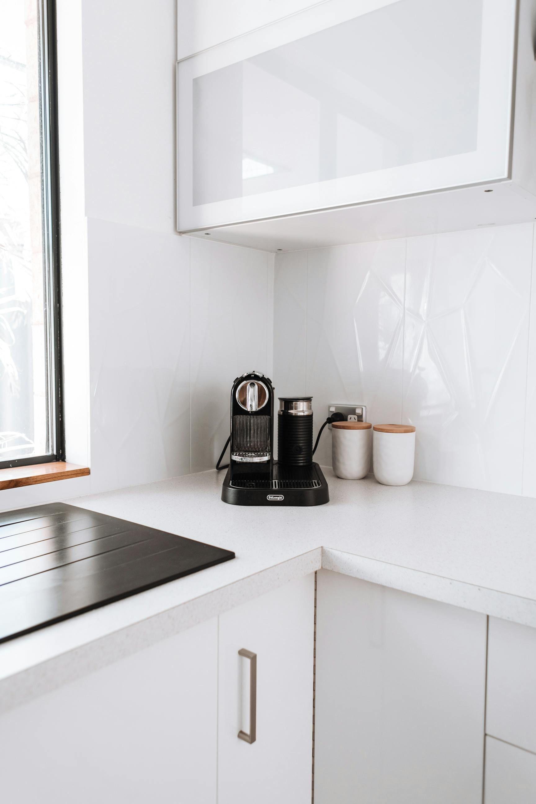 Moderne weiße Küche mit Dekton-Arbeitsplatte, schwarzer Kaffeemaschine und zwei weißen Behältern mit Holzdeckeln auf der Ecke der Arbeitsfläche.